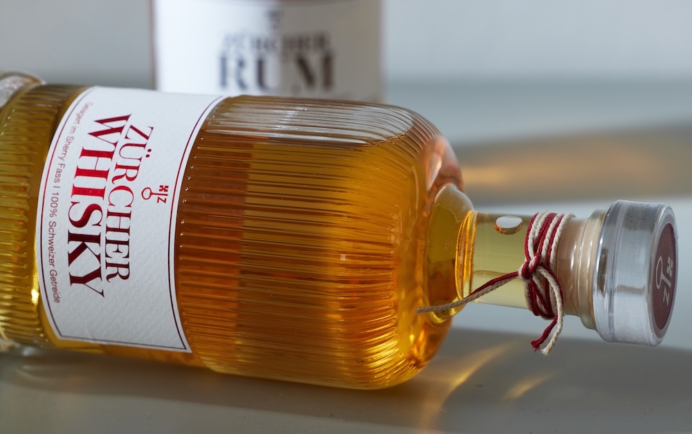 whisky flasche liegt seitlich, beschrifung in serifenschrift: 'zürcher whisky'ettikett: serifenschrift: 'zürcher whisky' in Rot veredelt, logo in der selben farbe darüber (schlüssel mit Z verbunden) 